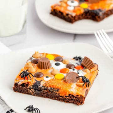 A slice of peanut butter Halloween eyeball brownies on a white plate with a glass of milk and another slice with a bite taken out of it in the background.