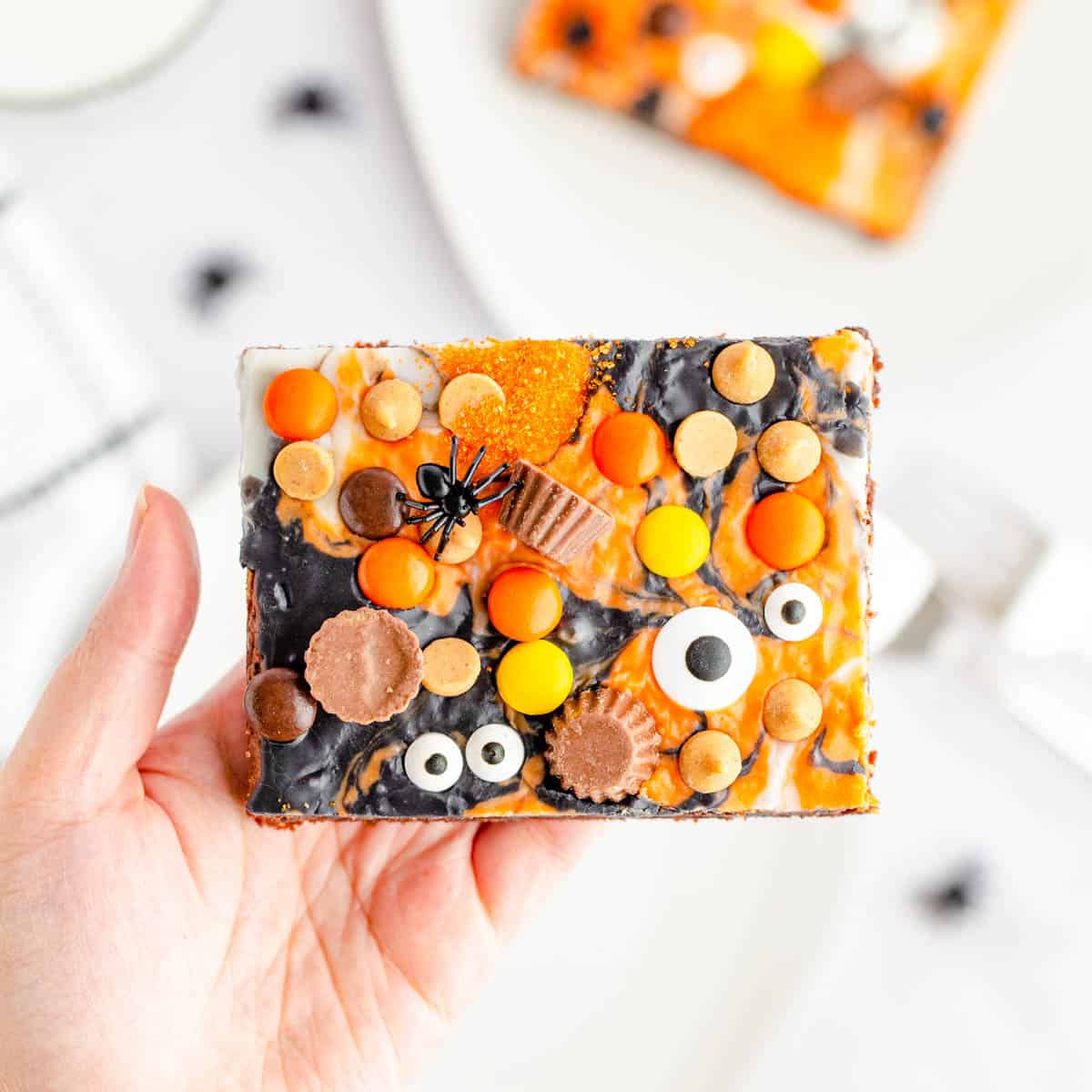 A hand holding a brownie so that you can see the top that is covered in black, orange, and white ganache along with mini peanut butter cups, peanut butter chips, Reese's pieces and candy eyeballs.
