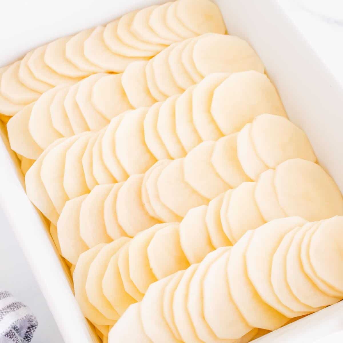 Russet potatoes that have been washed, peeled and sliced thinly and placed in a white casserole dish.