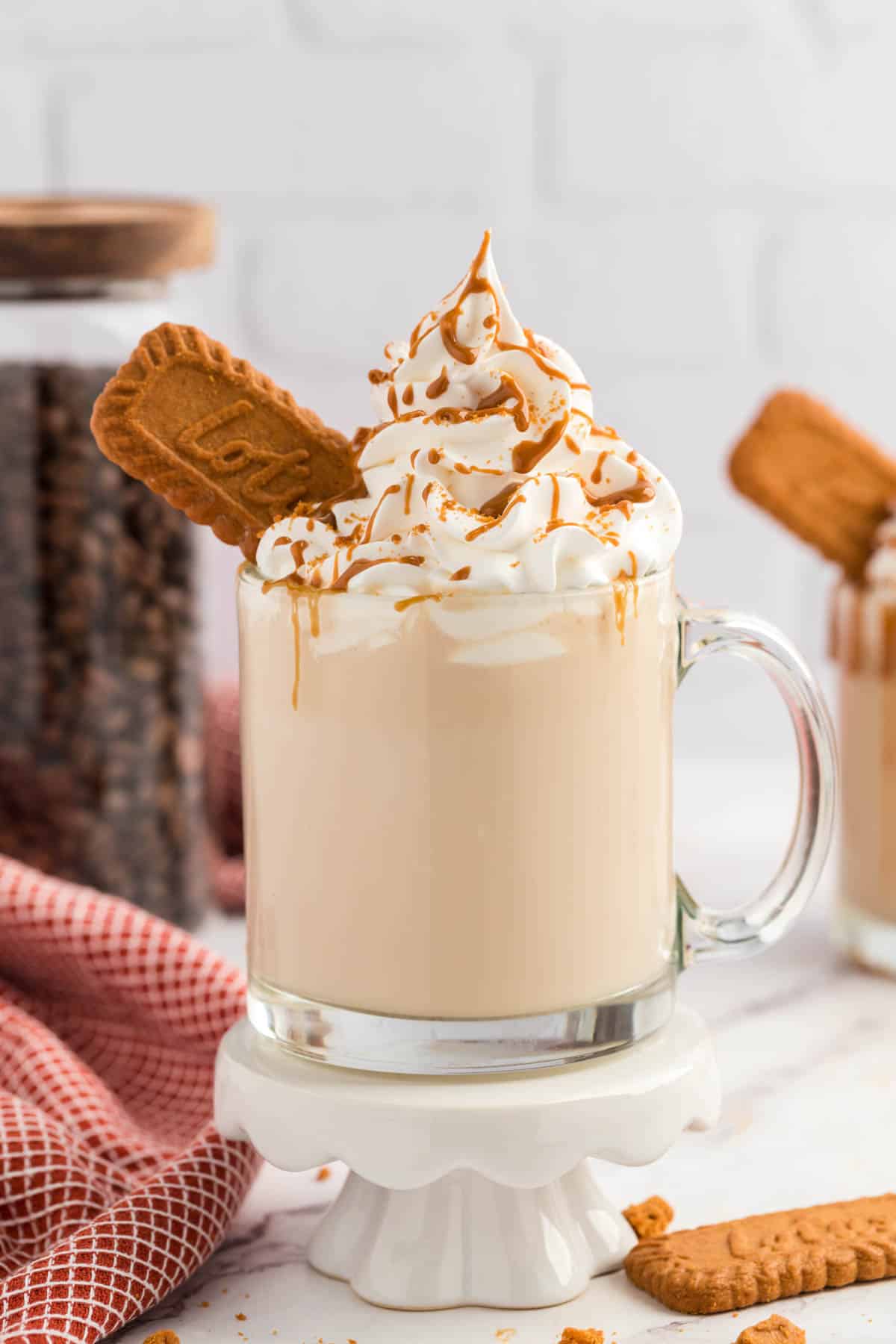 A glass coffee cup with a Biscoff Latte set on a white pedestal with scatter crumbs at the bottom.