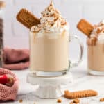 A glass coffee cup with a Biscoff Latte set on a white pedestal with scatter crumbs at the bottom.