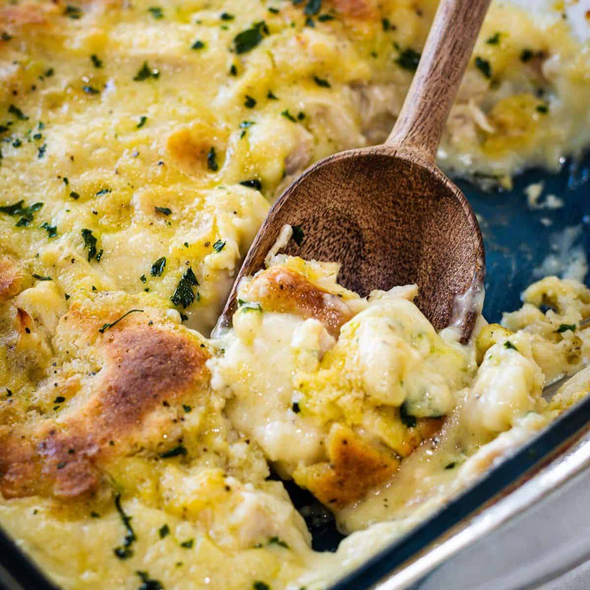 Chicken and dumplings in a clear casserole dish with a scoop being removed with a wooden spoon.