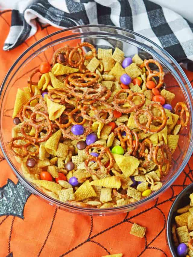 Overhead image of a clear bowl filled with Halloween Chex mix with a black and white checked tea towel in the background set on a orange and black placemat.