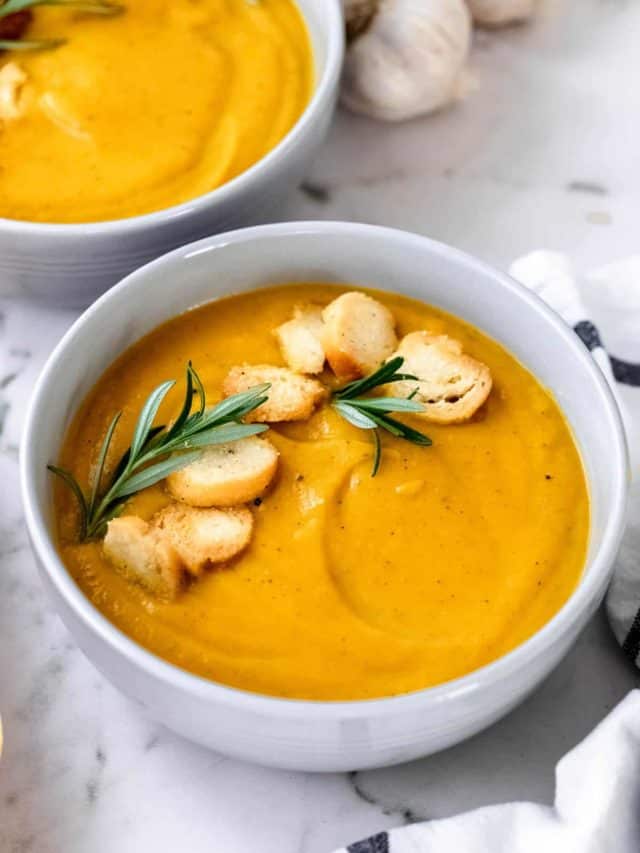 A white bowl filled with butternut squash soup garnished with rosemary and croutons.