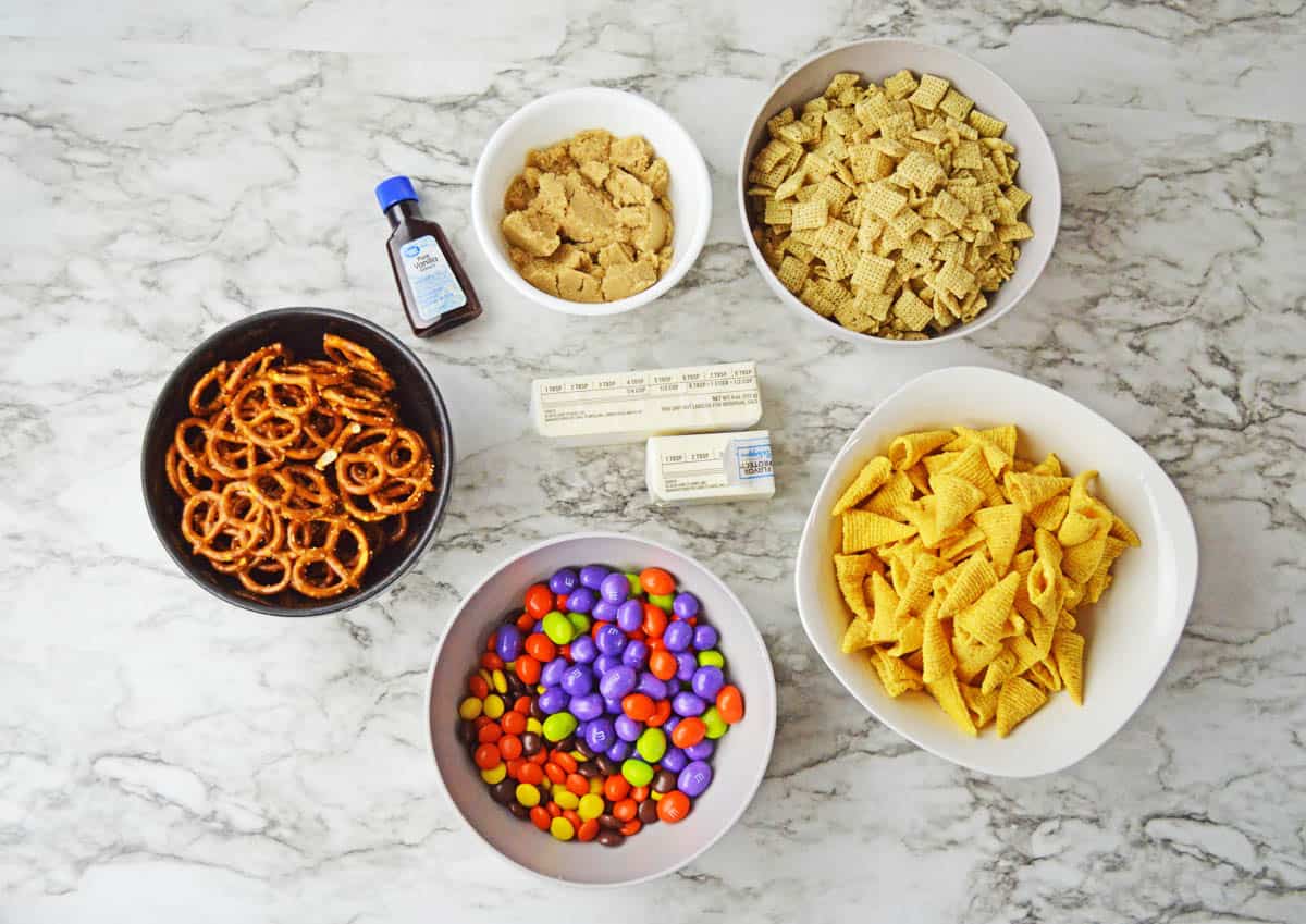 Image of ingredients needed to make Halloween Chex Mix; butter, brown sugar, vanilla, pretzels, rice chex, bugles, M&M's, and Reese's Pieces.