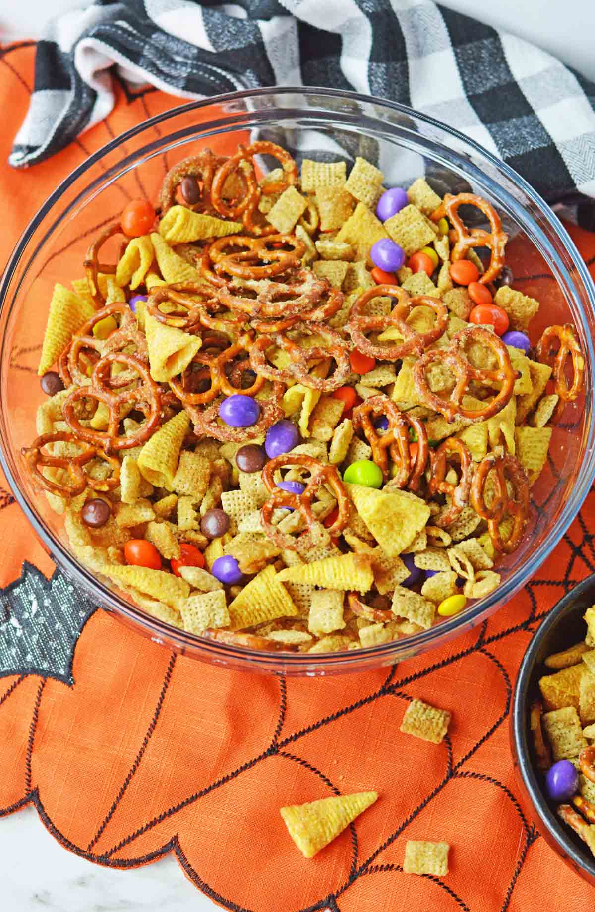 Overhead image of a clear bowl filled with Halloween Chex mix with a black and white checked tea towel in the background set on a orange and black placemat.