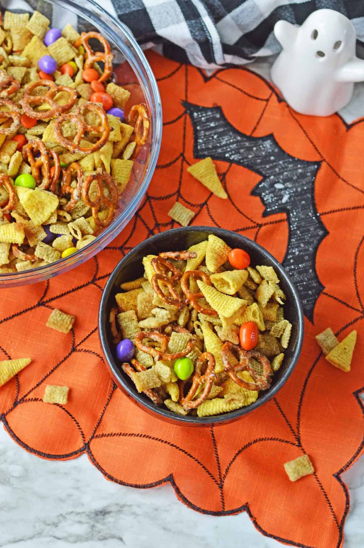 A small black bowl of chex mix along with a clear bowl full of the mix over to the side, all set on a festive halloween table.