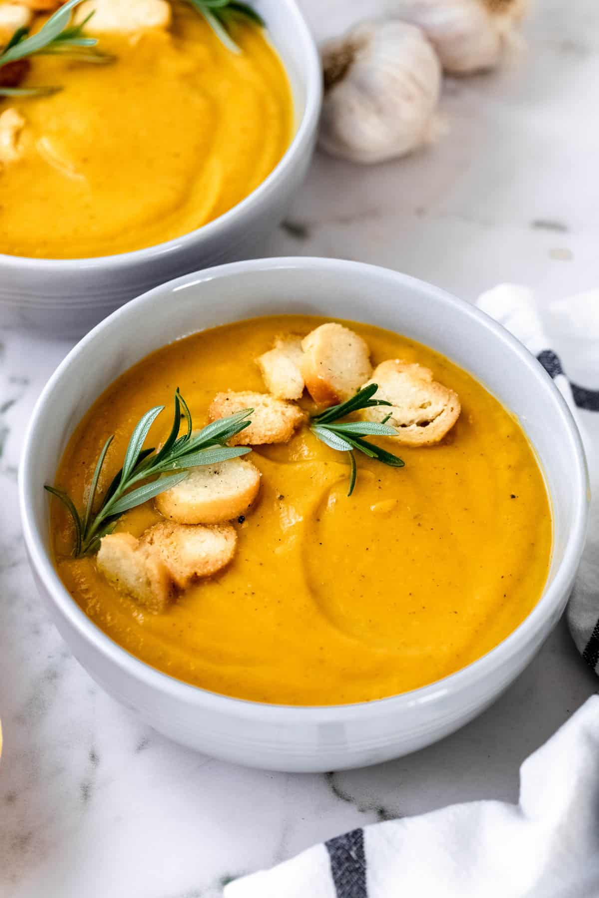 A white bowl filled with butternut squash soup garnished with rosemary and croutons.