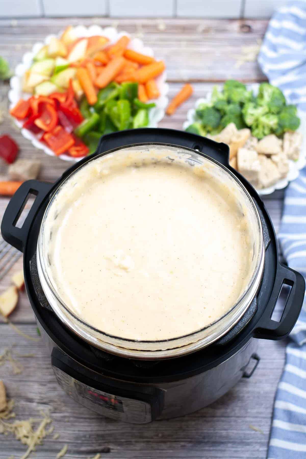 A Instant Pot filled with cheese fondue with a tray of raw veggies and bread cubes in the background.