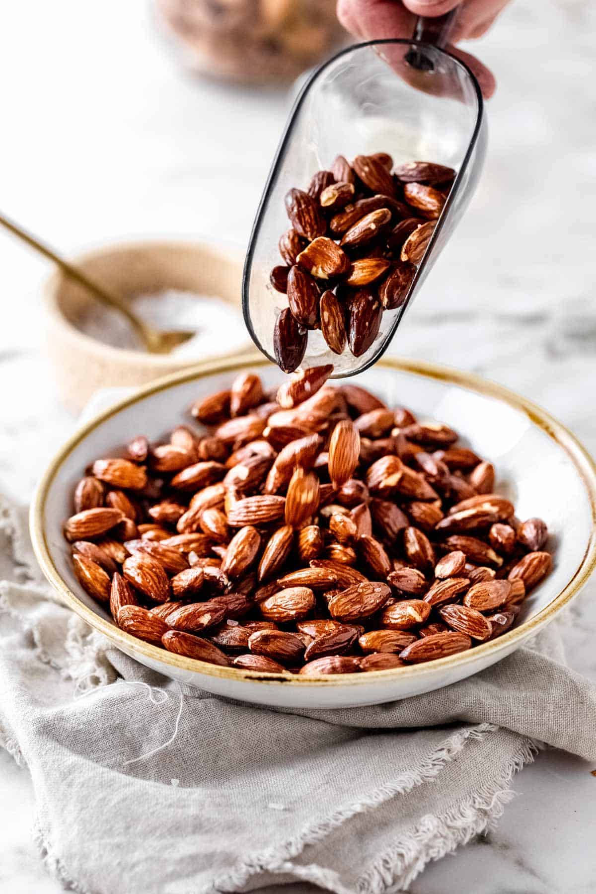 A scoop of roasted almonds being poured into a neutral bowl set on a beige cloth.