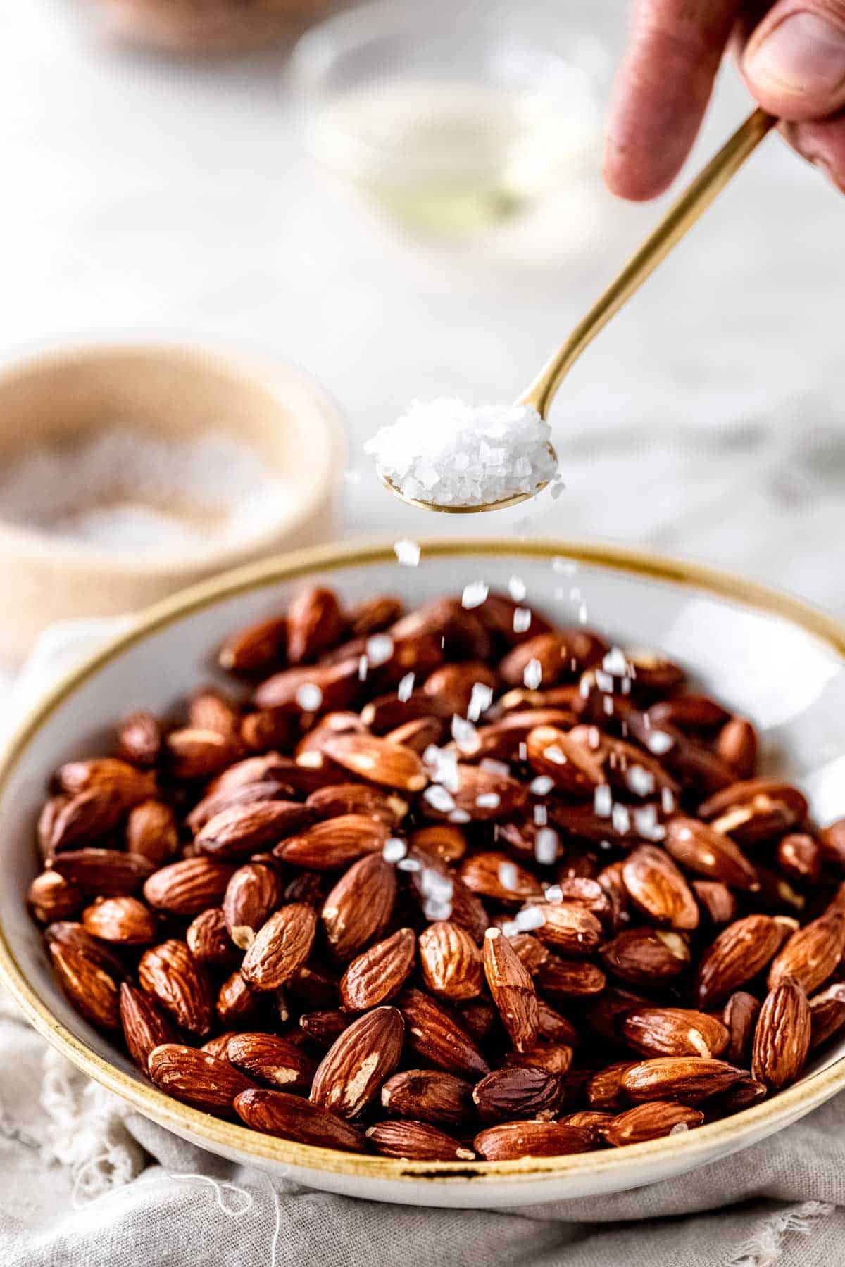 A bowl of roasted almonds with coarse salt being sprinkled onto them with a gold spoon.