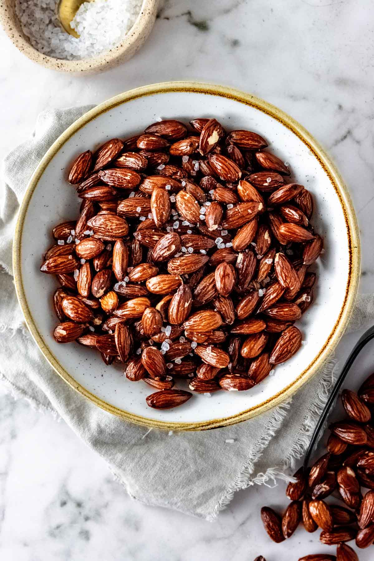 A white bowl with a gold rim filled with air fryer roasted almonds set on a neutral frayed napkin.