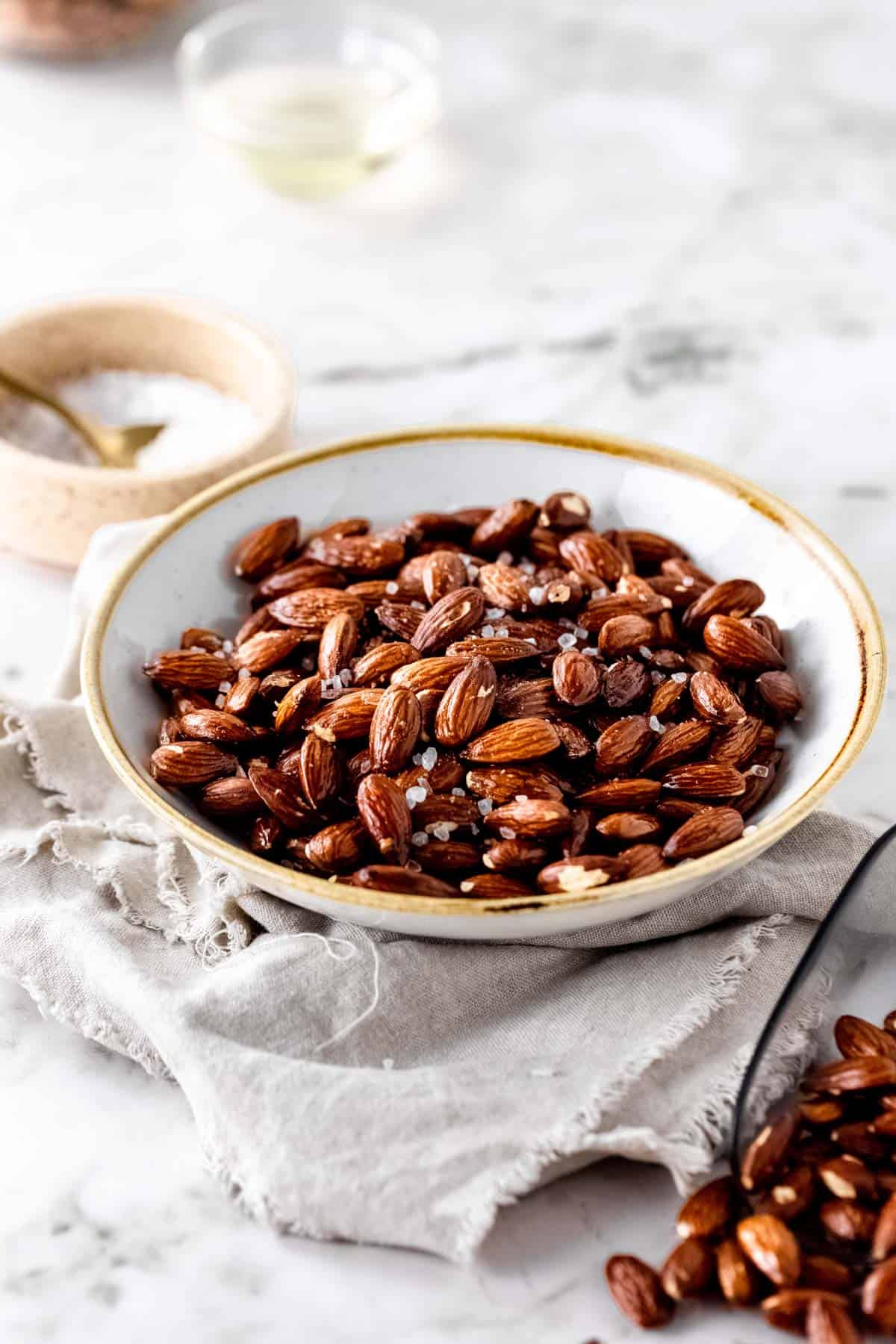 A bowl of air fryer roasted almonds sprinkled with set on a neutral napkin with a white bowl filled with coarse salt in the background.