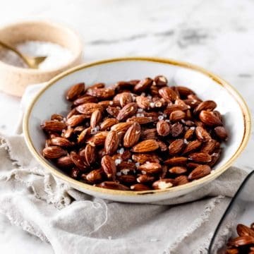 A bowl of air fryer roasted almonds sprinkled with set on a neutral napkin with a white bowl filled with coarse salt in the background.