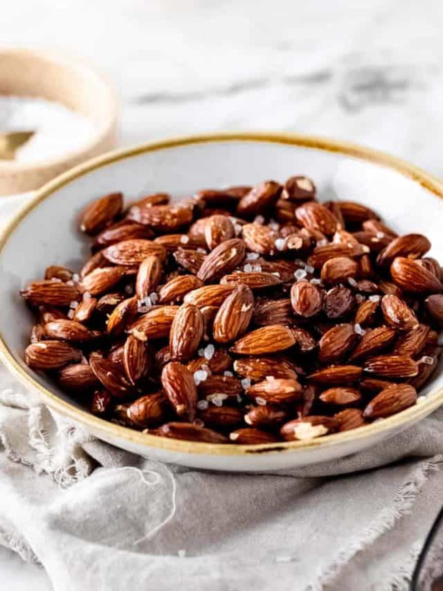 A bowl of air fryer roasted almonds sprinkled with set on a neutral napkin with a white bowl filled with coarse salt in the background.