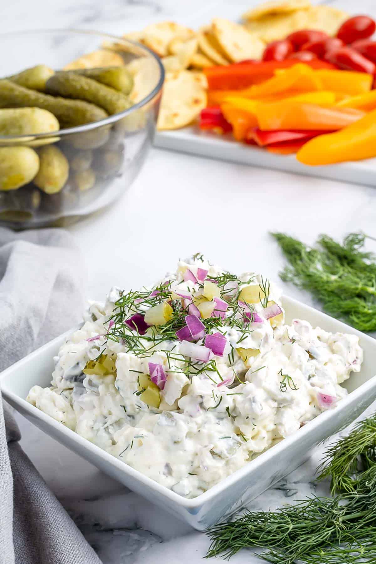 A bowl of dill pickle dip with fresh dill draped in front of the bowl and pickles, fresh veggies, and crackers in the background.