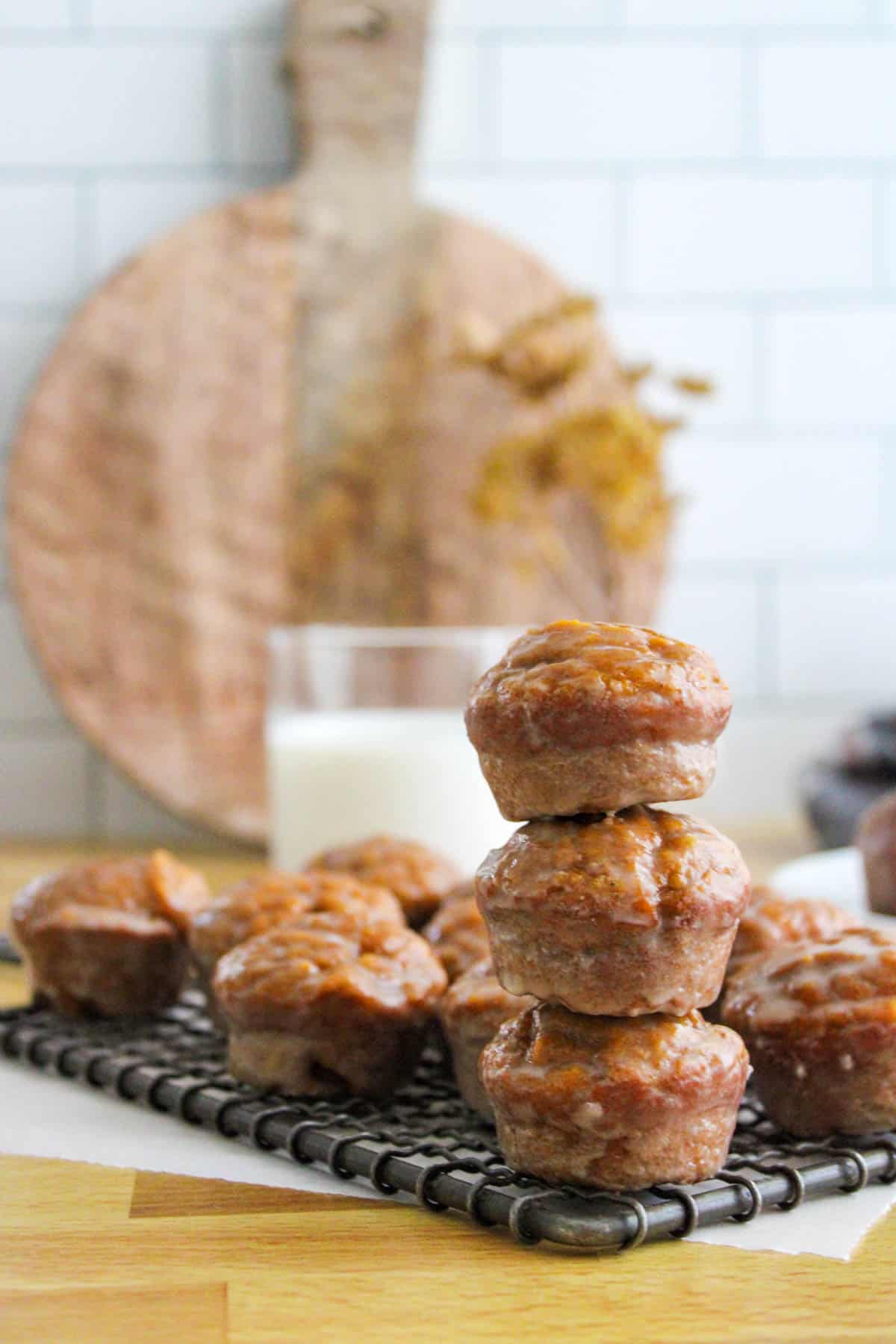 Glazed pumpkin donut muffins on a wire cooling rack stacked three high with muffins surrounding them and a glass of milk in the background.