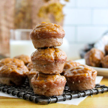 Glazed pumpkin donut muffins on a wire cooling rack stacked three high with muffins surrounding them and a glass of milk in the background.
