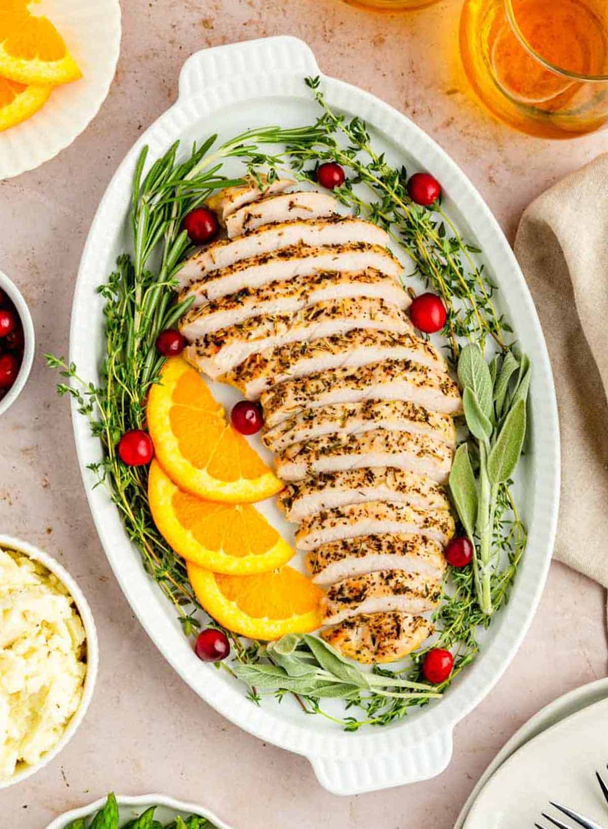Garlic herbed sliced turkey on a beautiful white platter garnished with fresh herbs, sliced oranges, and cranberries.