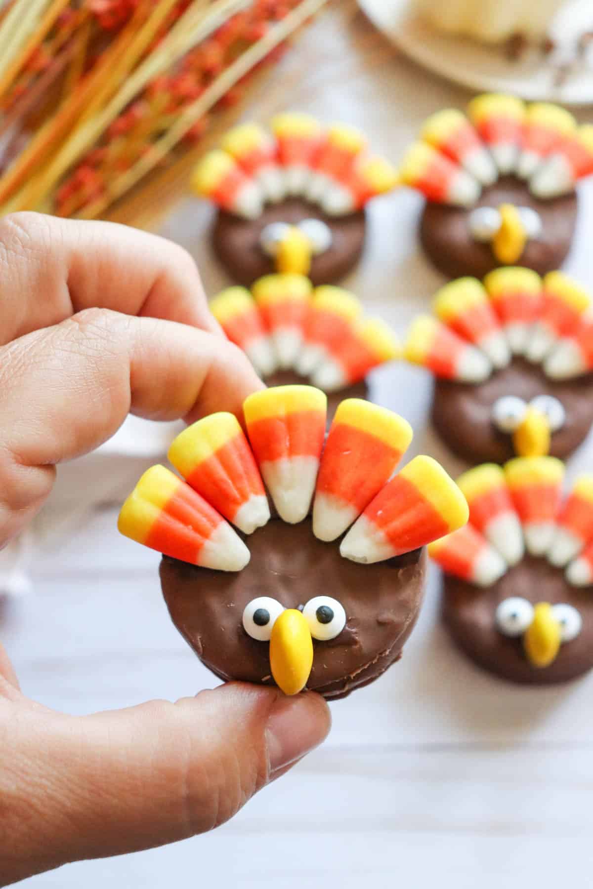 A hand holding a chocolate cookie decorated with candy corn, candy eyes and a yellow candy to look like a cute turkey.