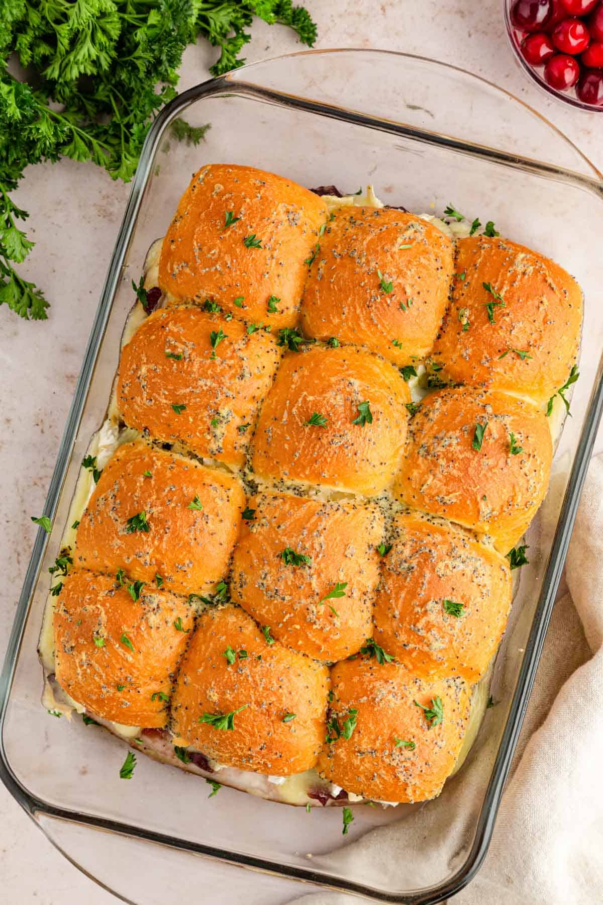 Overhead image of a glass baking dish filled with slider sandwiches and garnished with buttery poppy seed topping and parsley.