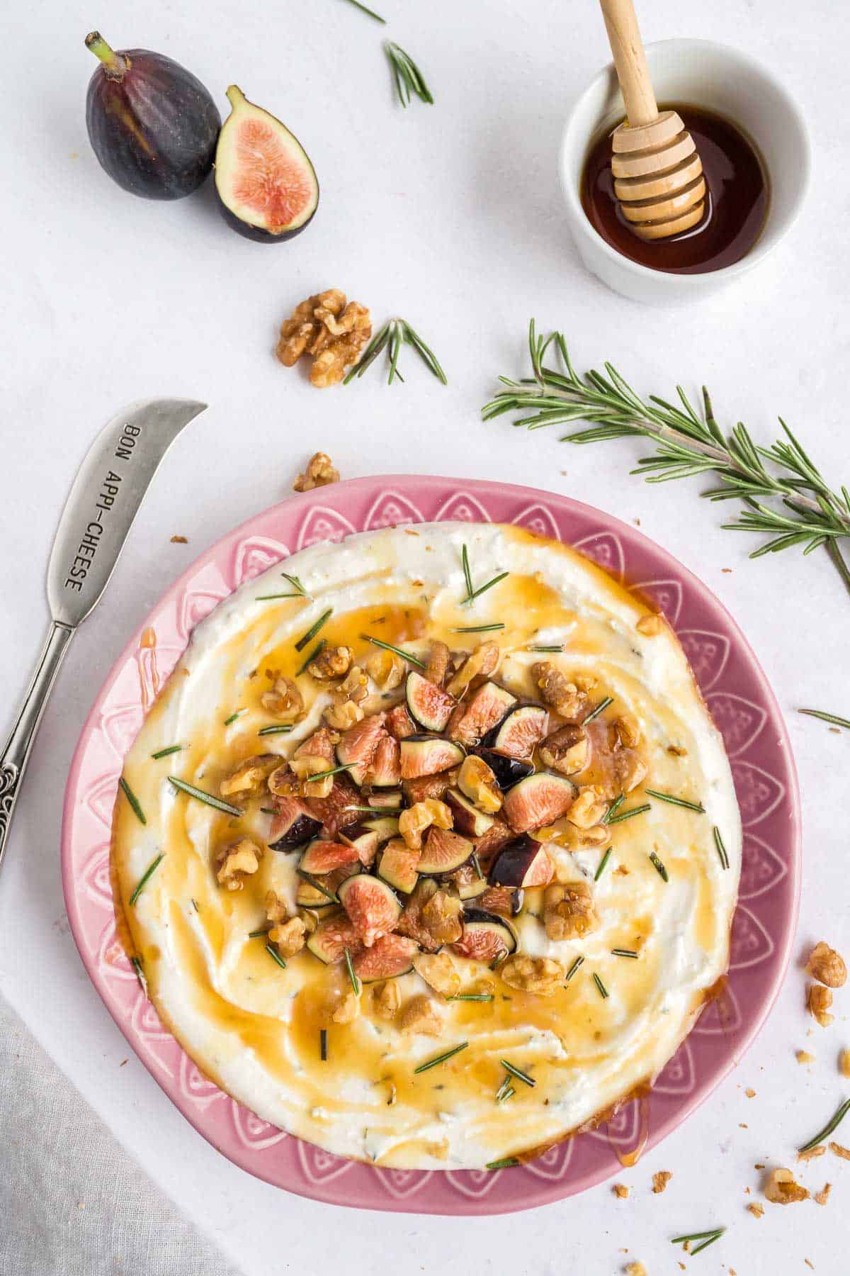 Whipped feta dip topped with nuts, figs, and drizzled with honey in a pink bowl on a neutral table with figs and a bowl of honey in the background.