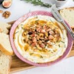 A pink bowl filled with a whipped feta dip with figs, nuts, and herbs with a slice of dipped bread on the bowl.