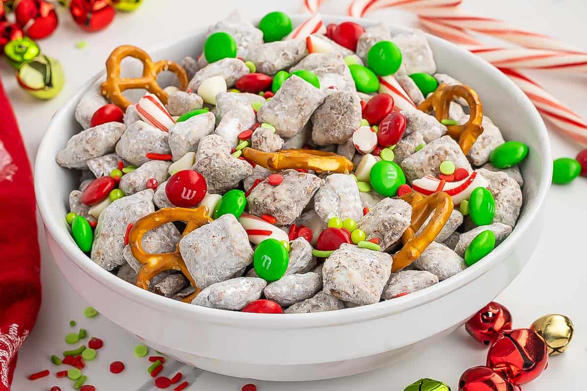 A white bowl with muddy buddies filled with Christmas m&ms, pretzels, candy canes, and white chocolate chips.