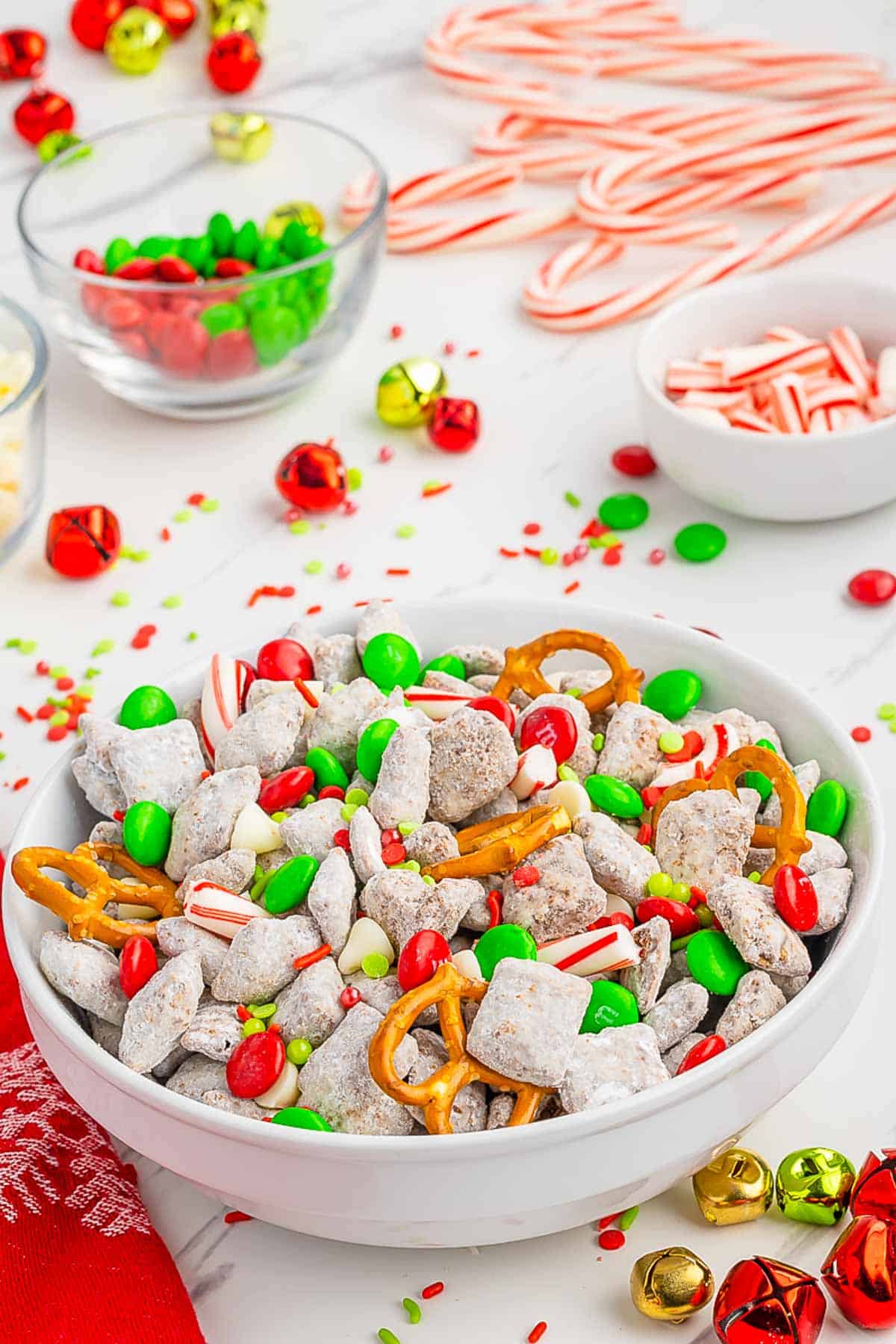A white bowl filled with Christmas Muddy Buddy Chex Mix set on a festive table with scattered sprinkles, christmas bells and candy canes.