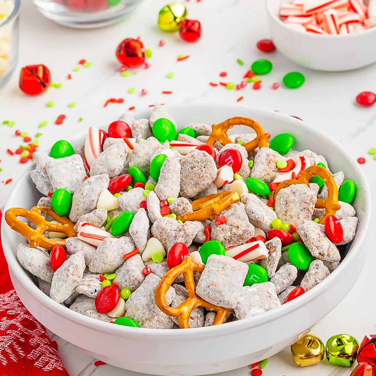 A white bowl of Christmas Muddy Buddies on a white wood table with Holiday decor.