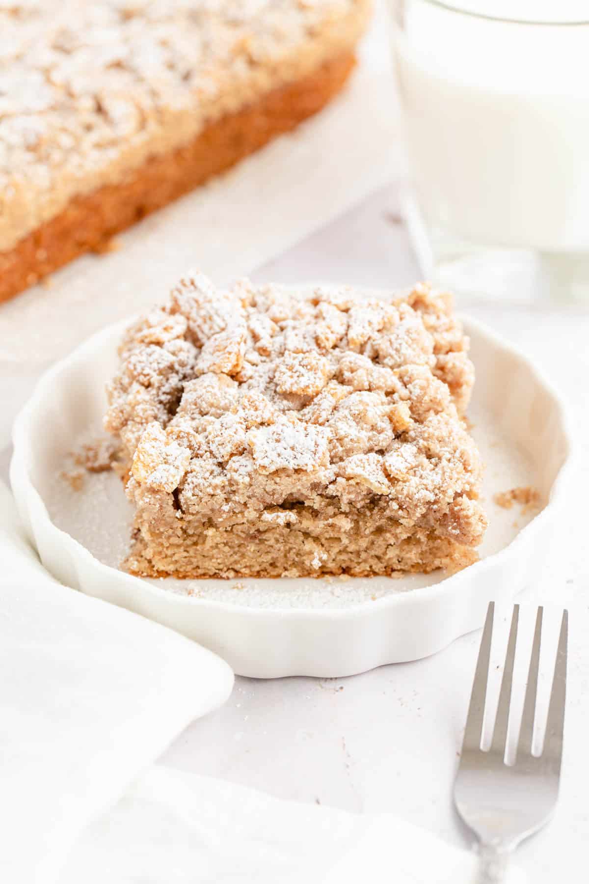 A slice of cinnamon toast crunch cake on a white plate with the whole cake in the background.