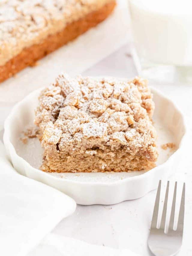 A slice of cinnamon toast crunch cake on a white plate with the whole cake in the background.
