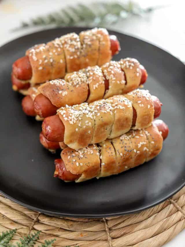 A black plate stacked with keto pretzel dogs topped with sesame and poppy seeds.