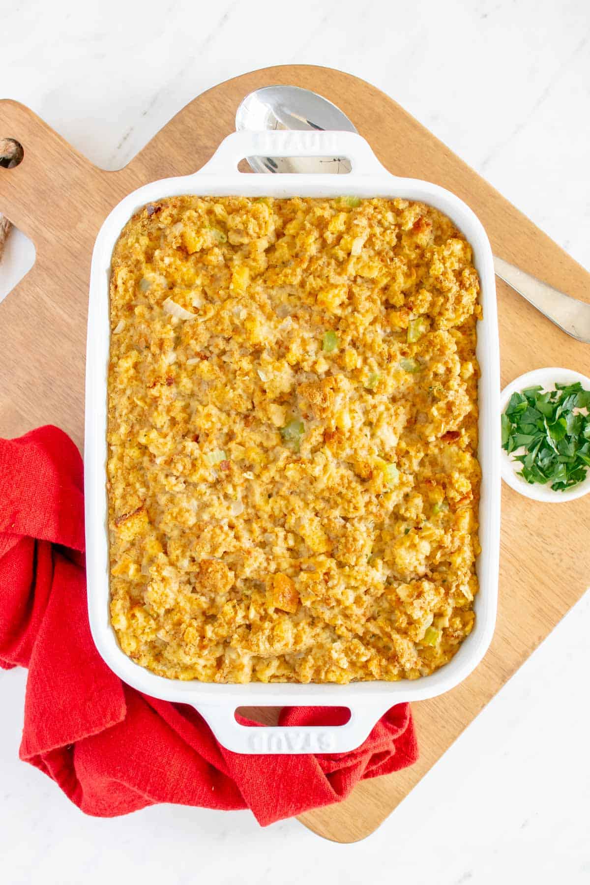 A white baking pan of stuffing on a wooden board with a red tea towel underneath and cut parsley in a bowl to the side.