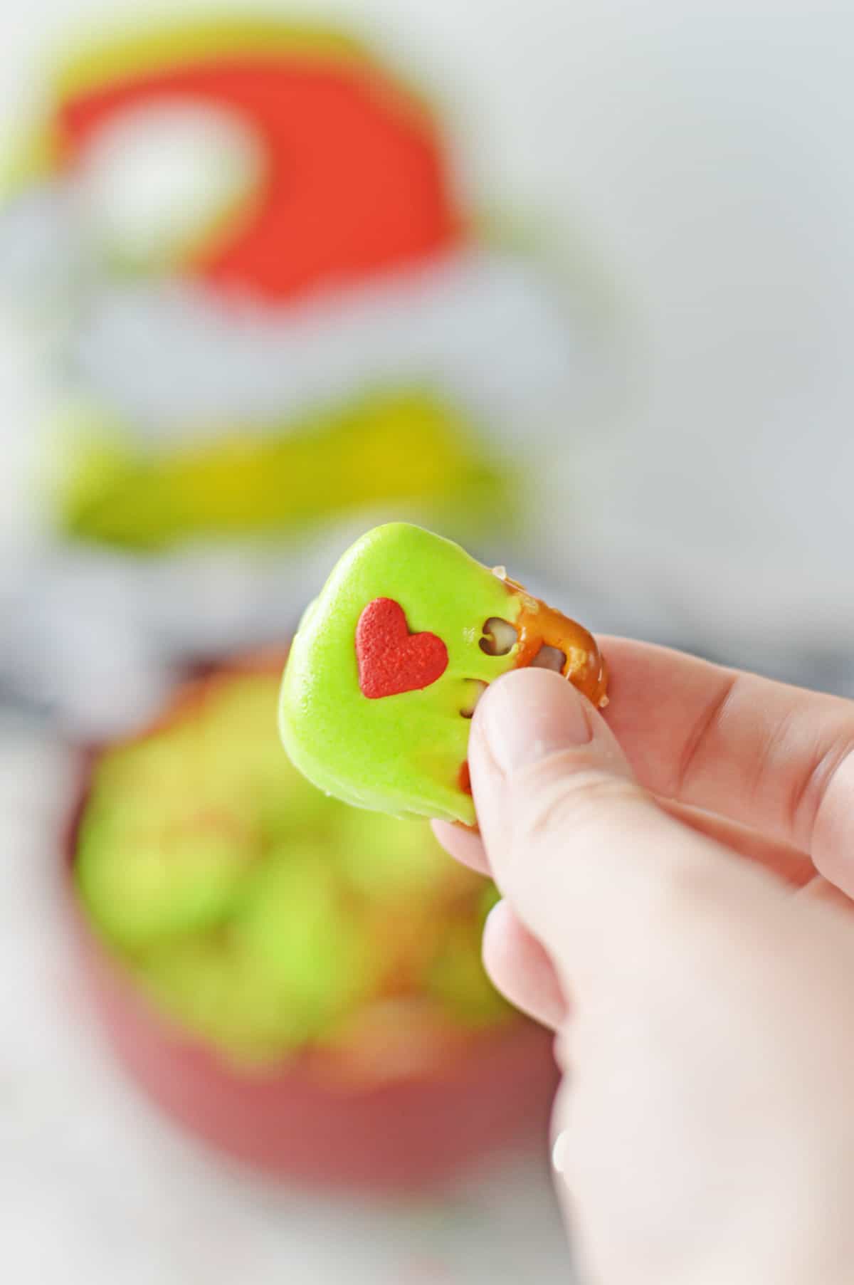 A hand holding a dipped Grinch Pretzel Bite with a heart candy on the top.
