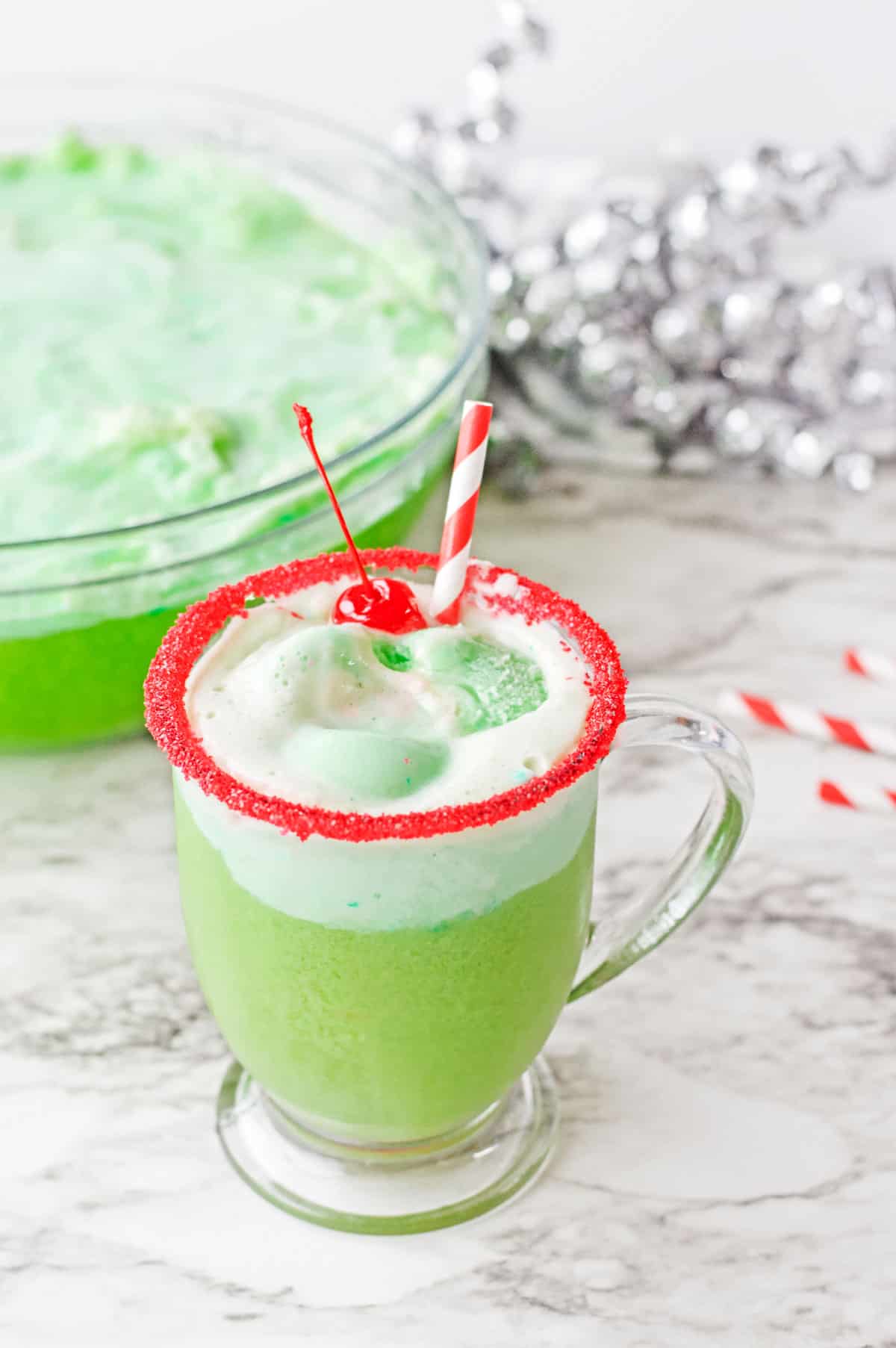 A glass of grinch punch with the punch bowl and a silver confetti streamer in the background.