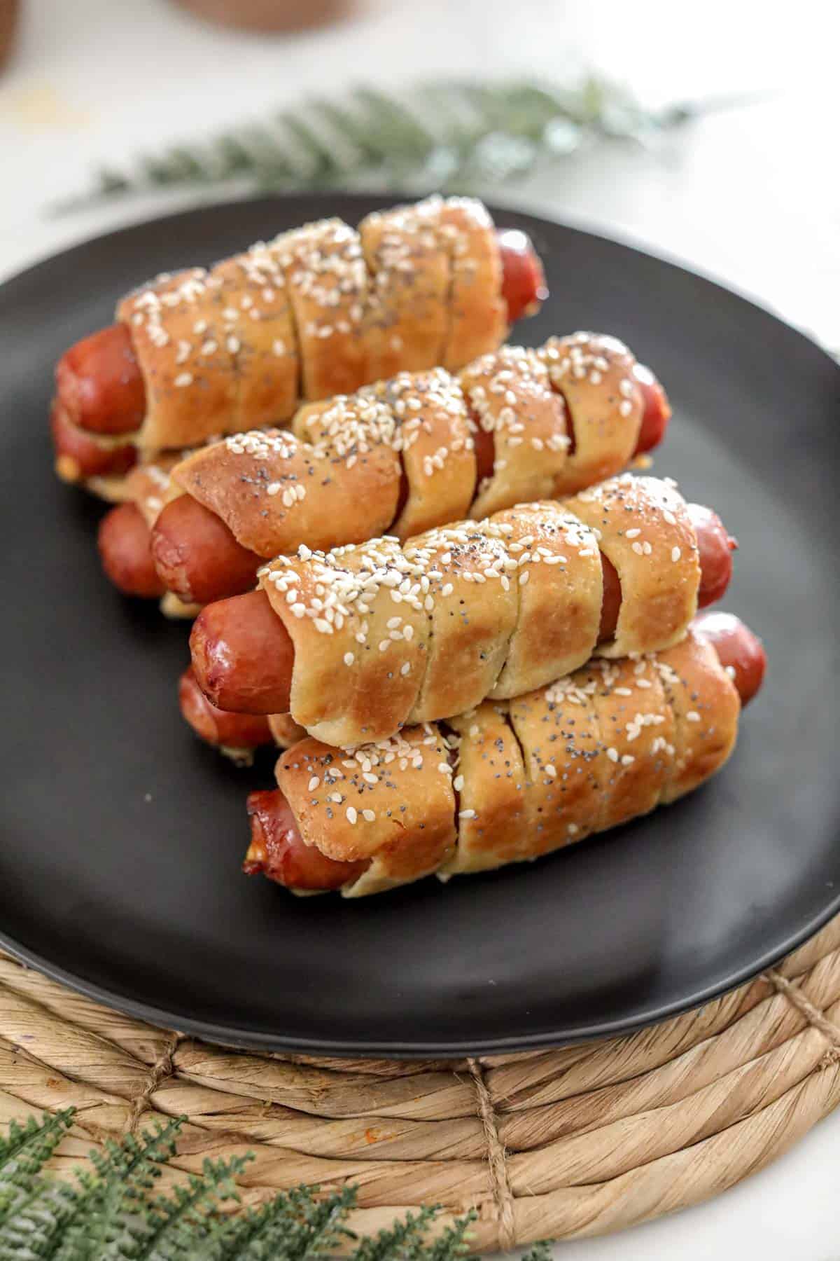 A black plate stacked with keto pretzel dogs topped with sesame and poppy seeds.