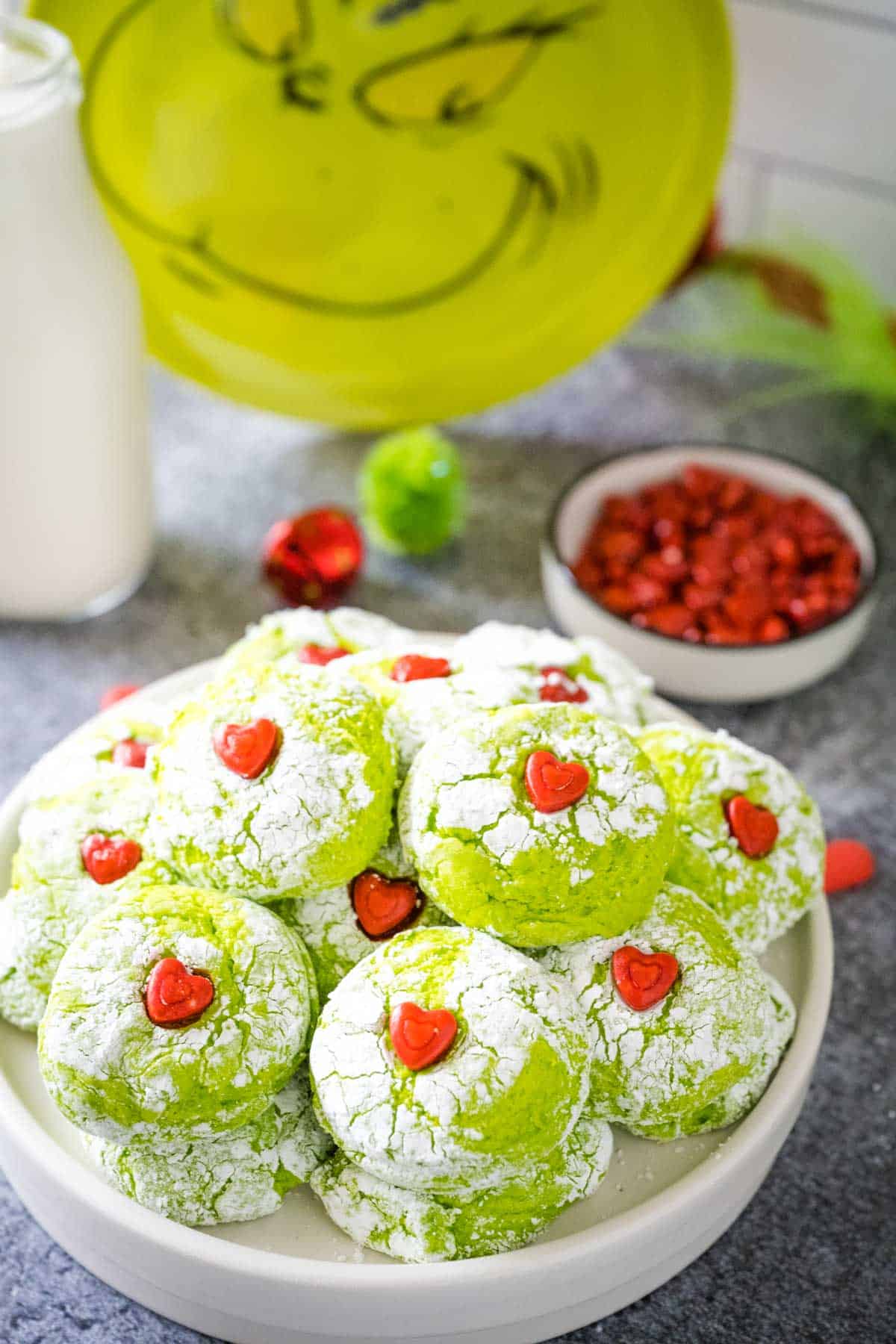 A white plate filled with grinch cookies with a small bowl of candy hearts and the face of the grinch in the background.