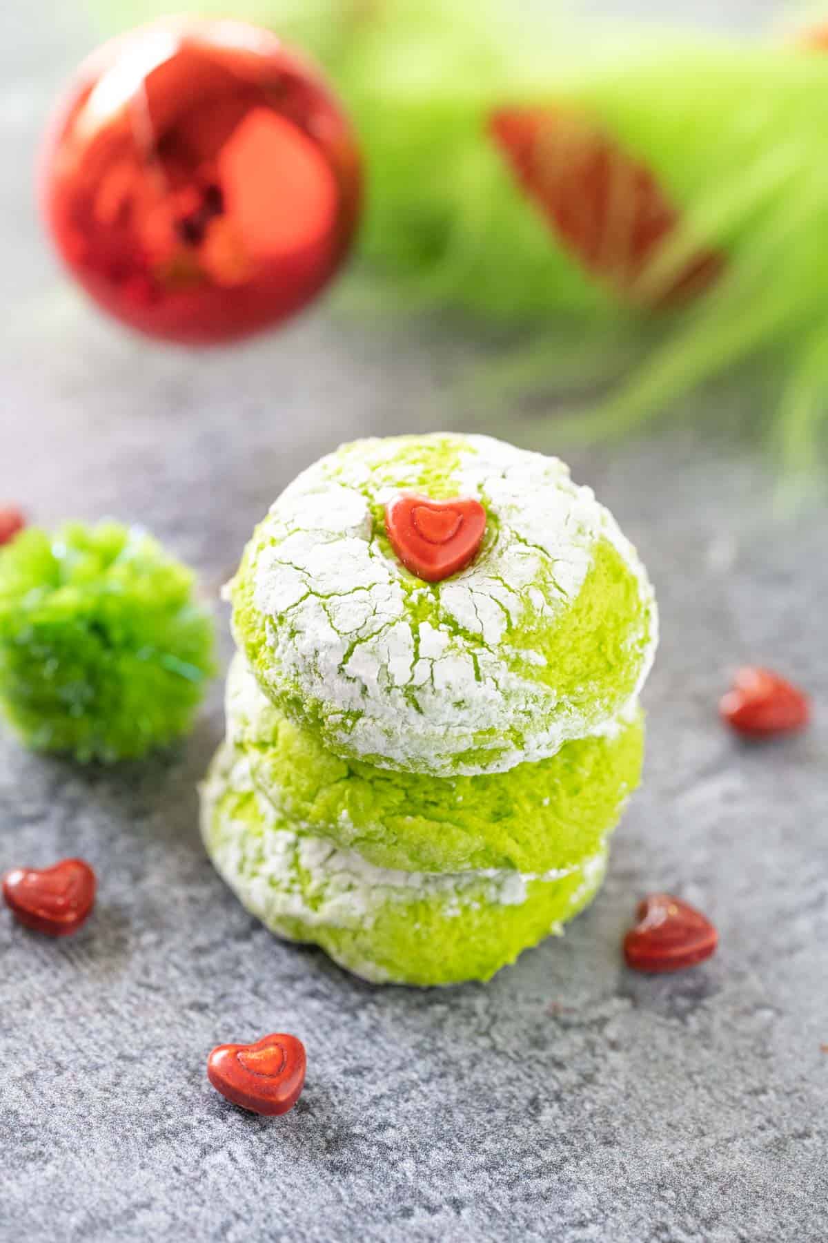 Three Grinch cookies stacked on a grey surface with hearts scattered and fuzzy a grinch tail in the background.