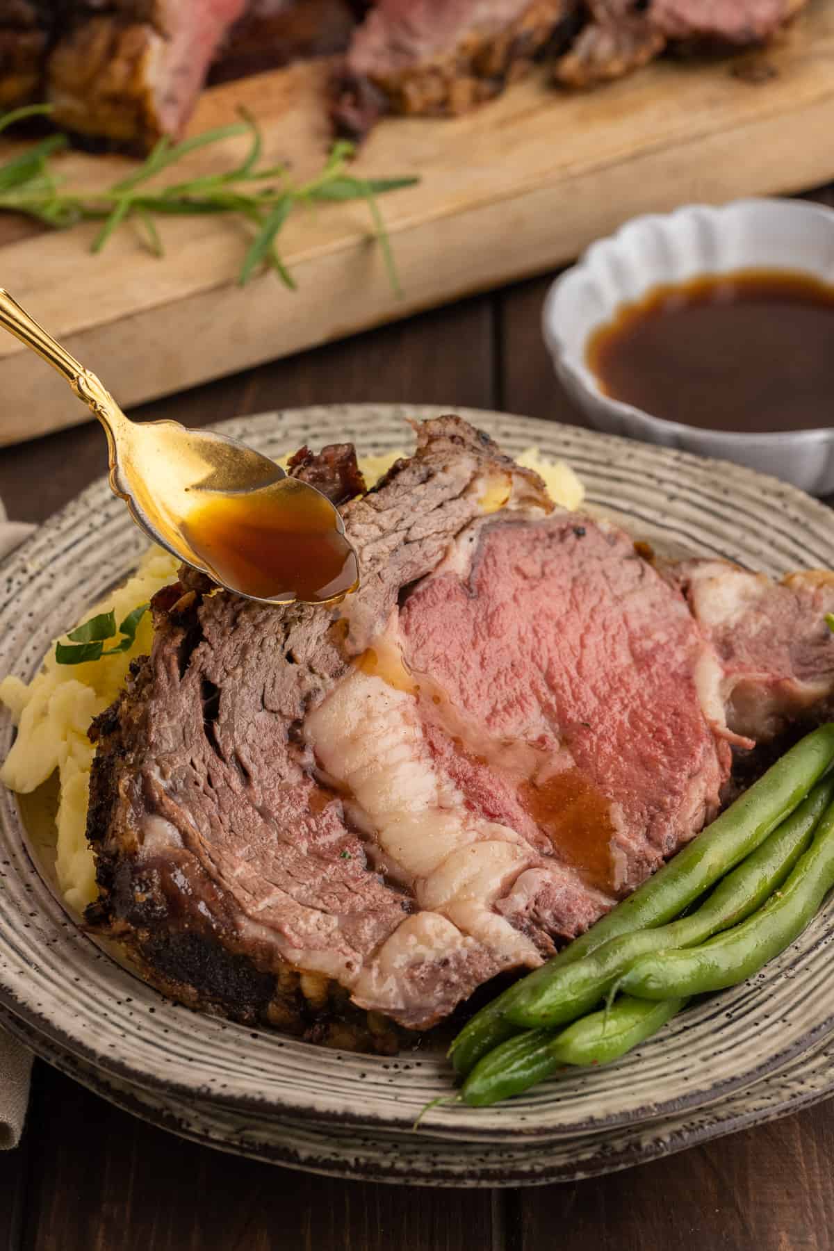 Prime rib on a plate with fresh green beans and mashed potatoes with au jus being drizzled over the meat.