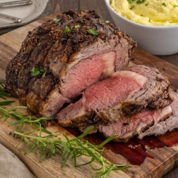 A sliced prime rib garnished with rosemary on a cutting board with a bowl of creamy mashed potatoes in the background.