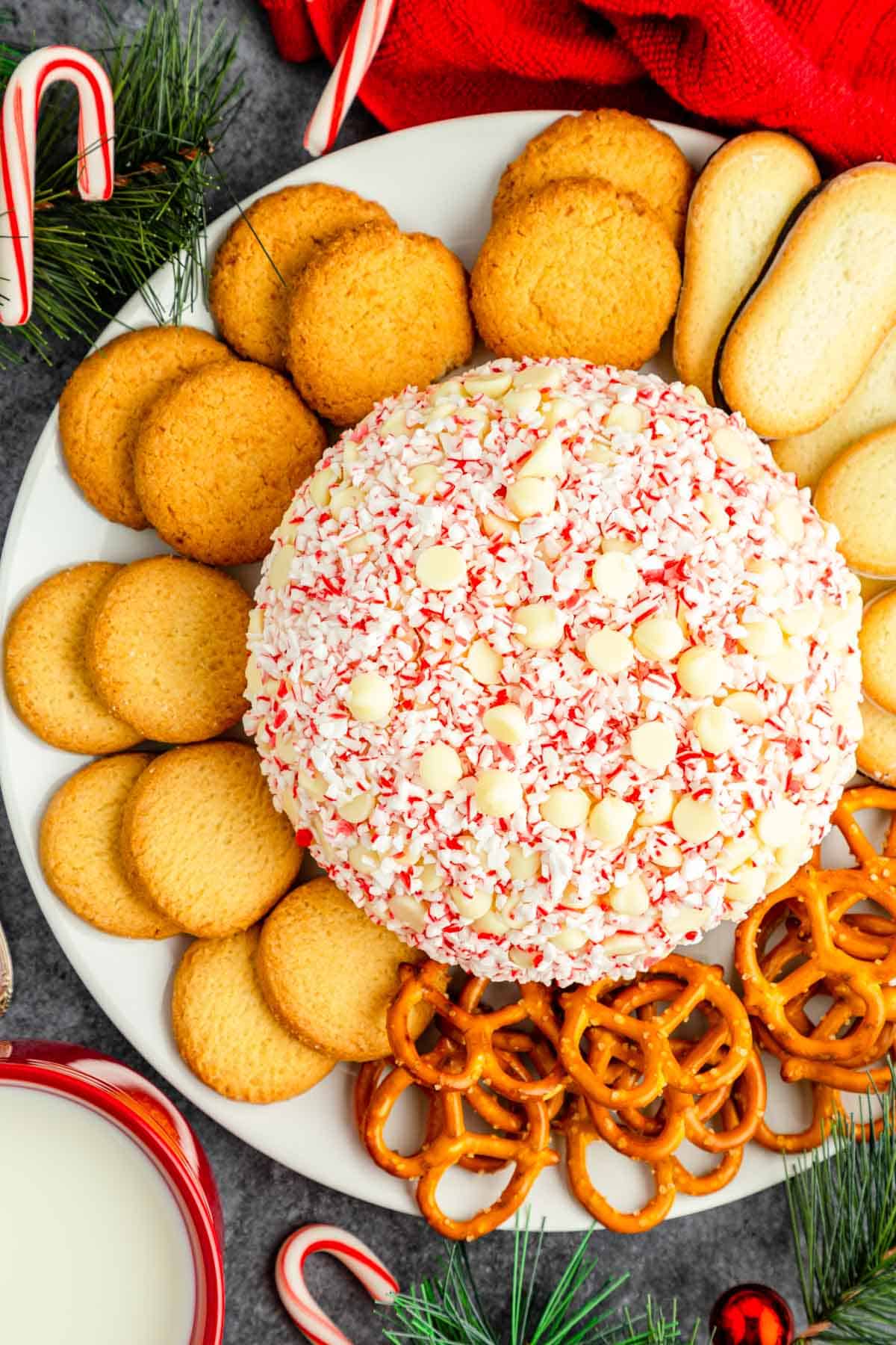 An overhead shot of a white plate with white chocolate peppermint cheesecake ball surrounded by cookies and pretzels on a grey table with evergreens and candy canes.