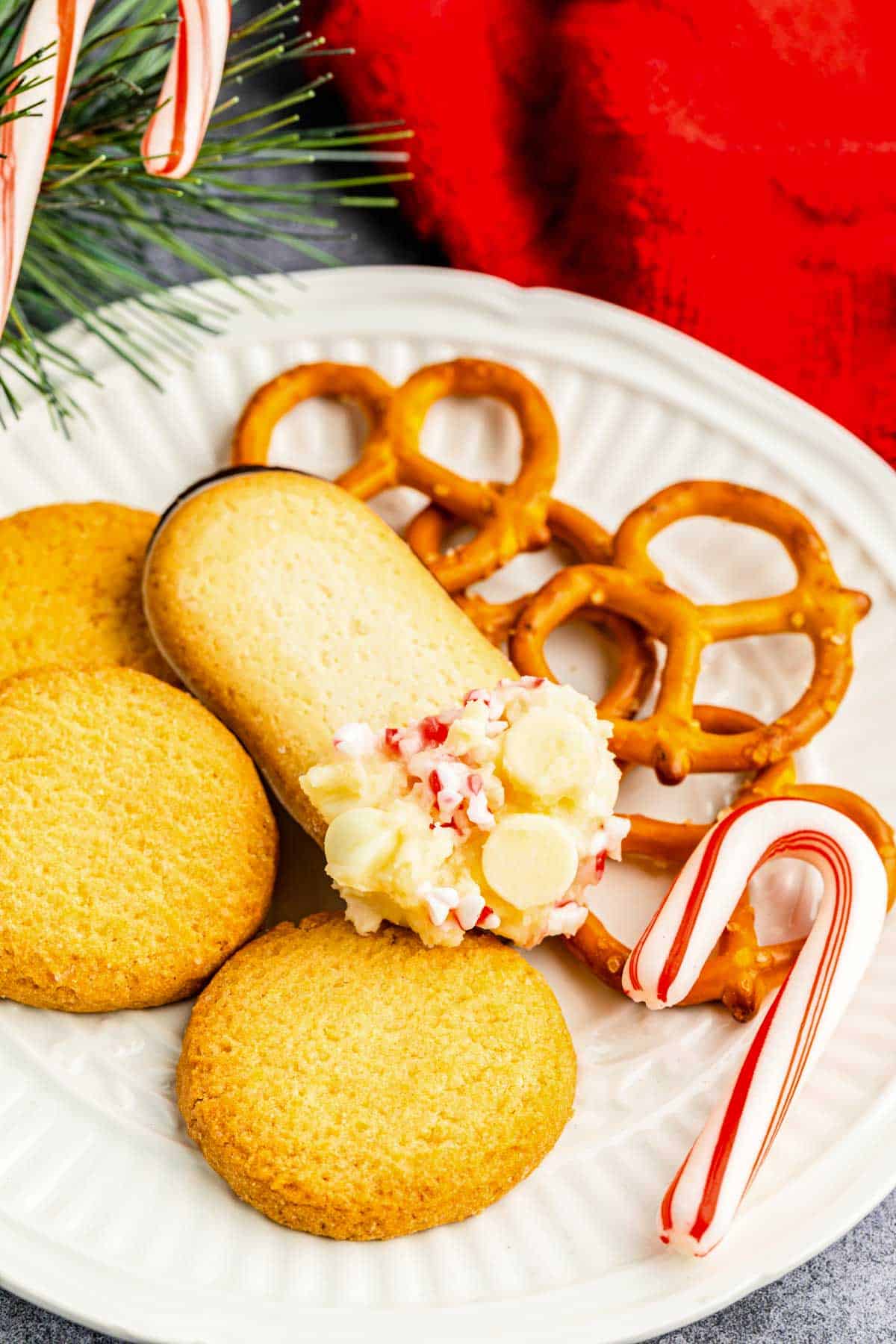 A white plate with assorted cookies, pretzels and a candy cane topped with peppermint cheesecake dip.
