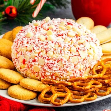 A white chocolate peppermint cheesecake ball on a plate with pretzels and cookies decorated for the holidays.
