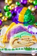 A slice of Mardi Gras King Cake on a white plate with red, gold, and purple beads scattered.