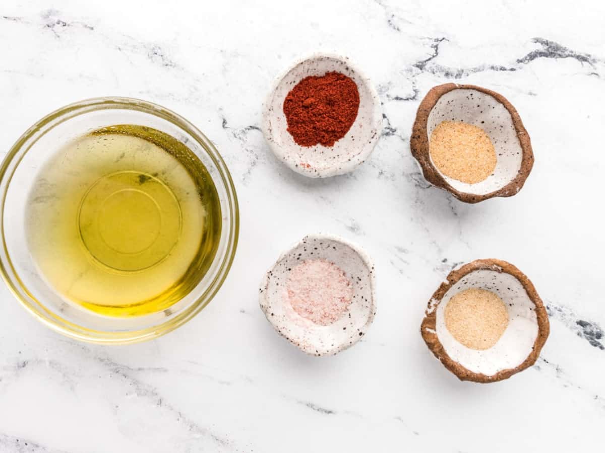 Bowls of olive oil, paprika, garlic powder, salt, and onion powder on a marble table.