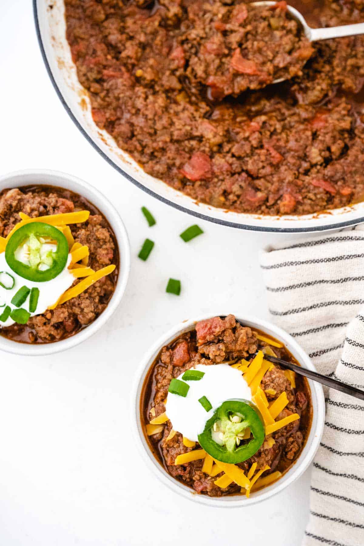 An overhead shot of two bowls of chili garnished with sour cream, cheese, and chives, and a pot of chili on a white table.