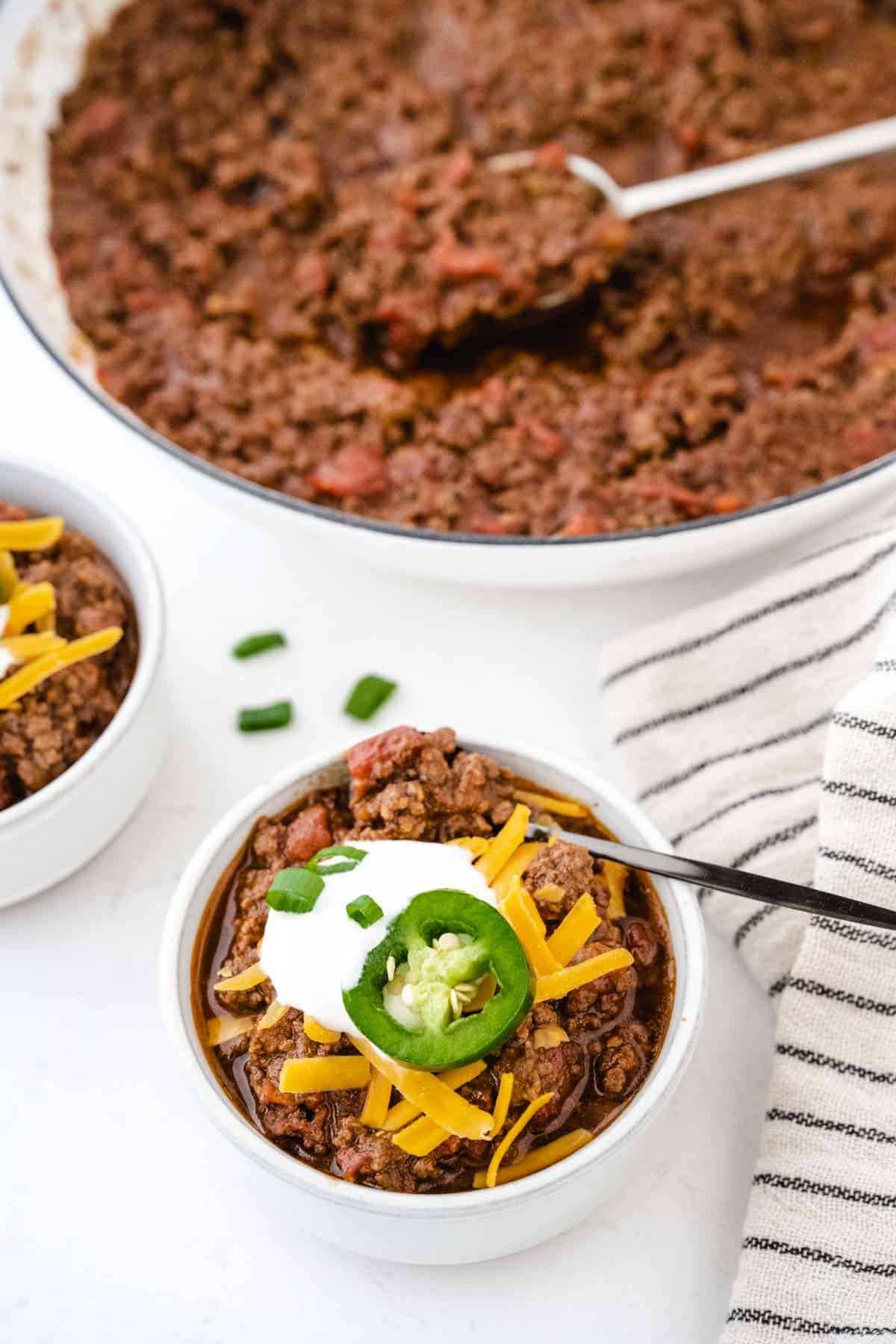 A bowl of chili with all the fixings with a pot of chili in the background.