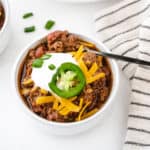 A bowl of no bean chili topped with shredded cheddar cheese, a dollop of sour cream, a slice of jalapeno, and green onions.