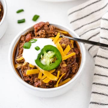 A bowl of no bean chili topped with shredded cheddar cheese, a dollop of sour cream, a slice of jalapeno, and green onions.